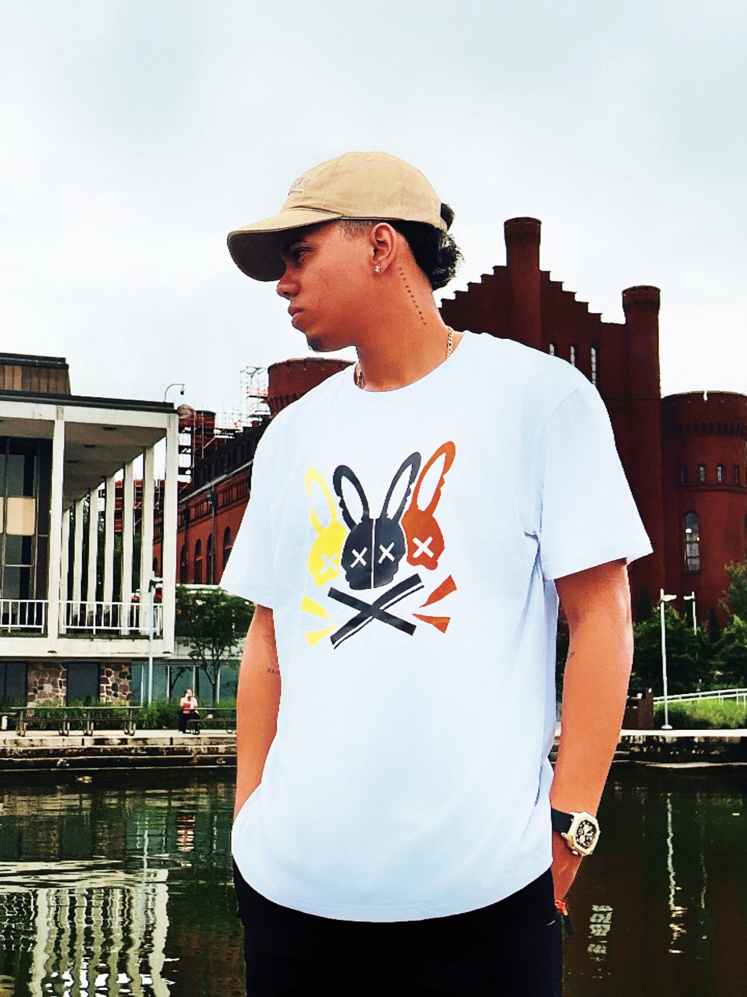 Person wearing a white t-shirt with a graphic design, standing by a waterfront with buildings in the background.
