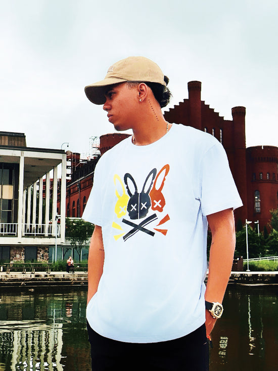 Person wearing a white t-shirt with a graphic design, standing by a waterfront with buildings in the background.