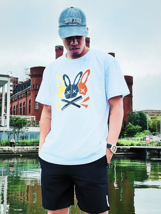 Person wearing a light blue t-shirt with a graphic design and a cap, standing by a waterfront.